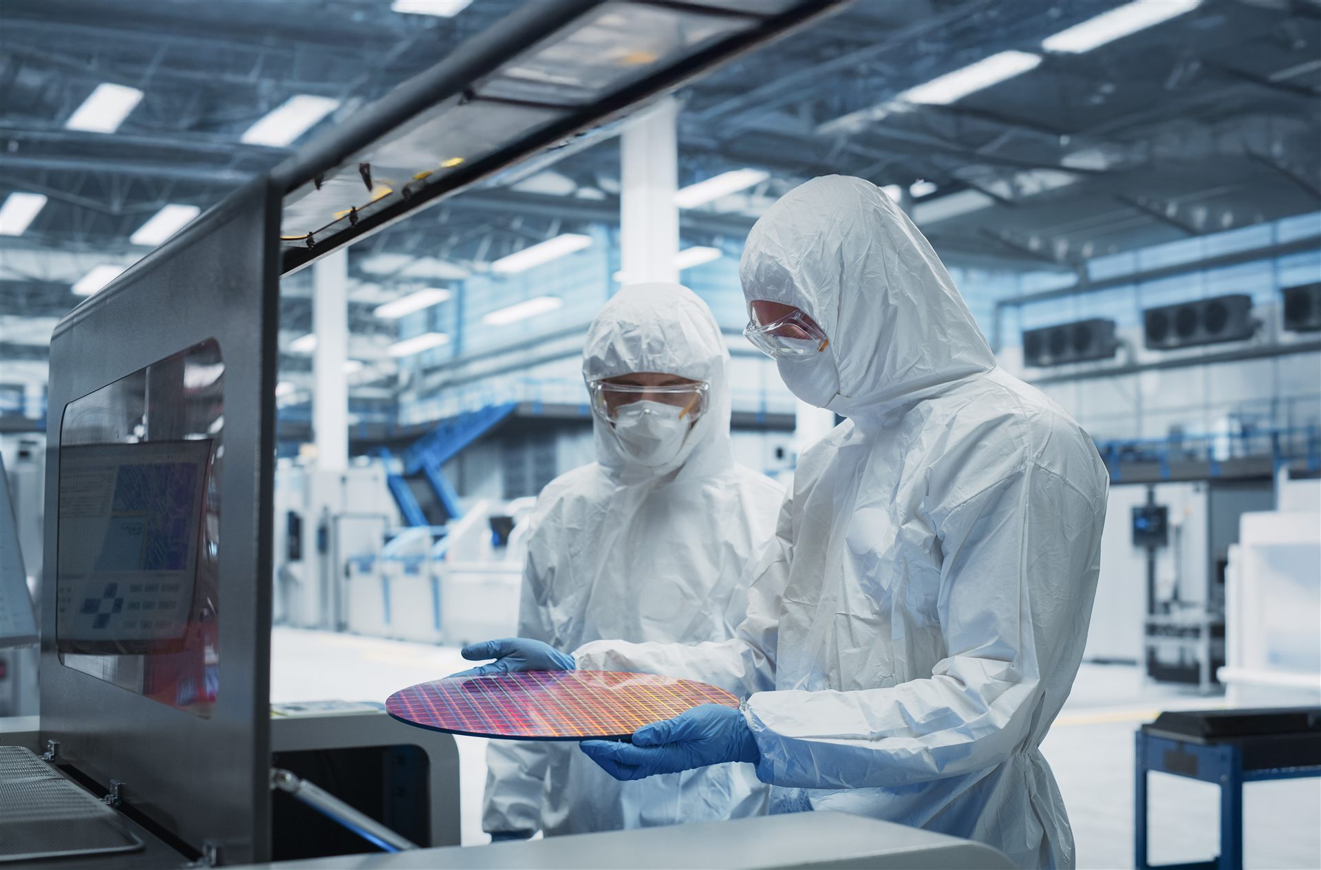 Zwei Personen in Reinraumanzügen begutachten einen Silizium-Wafer in einer Halbleiter-Reinraumfertigung.